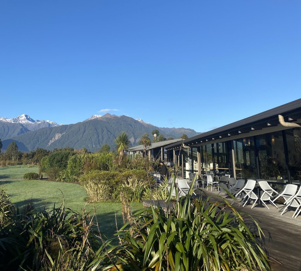 A photo of a cafe with brown beams on the outside and a full glass front, under beautiful blue skies. The views in the background are of Mount Cook and the Southern Alps of New Zealand.