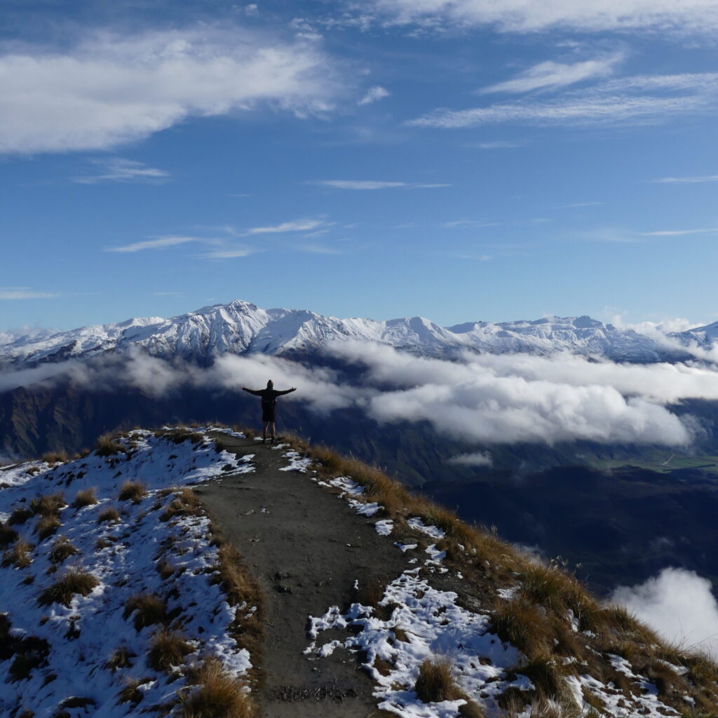 A small figure stands at the top of a mountain with his arms stretched wide over an epic, snowy view.