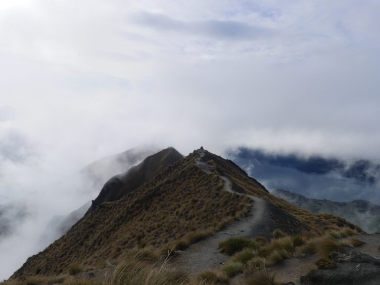 A winding path leading over towards the edge of a small grassy hill with a small patch of lake Wānaka visible below but mainly obstructed by clouds and only a small bit of sky poking through