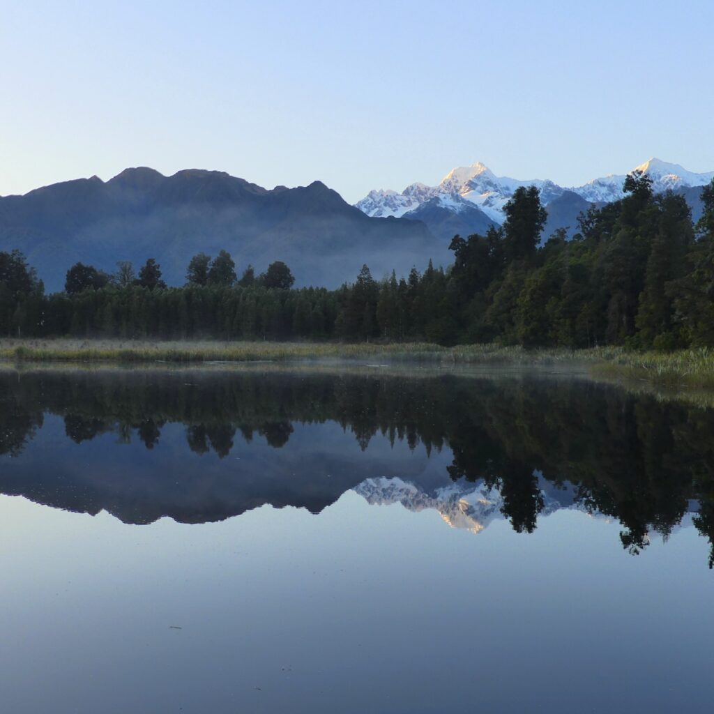 A lake reflecting the surrounding trees and snow capped mountains perfectly upon its surface.