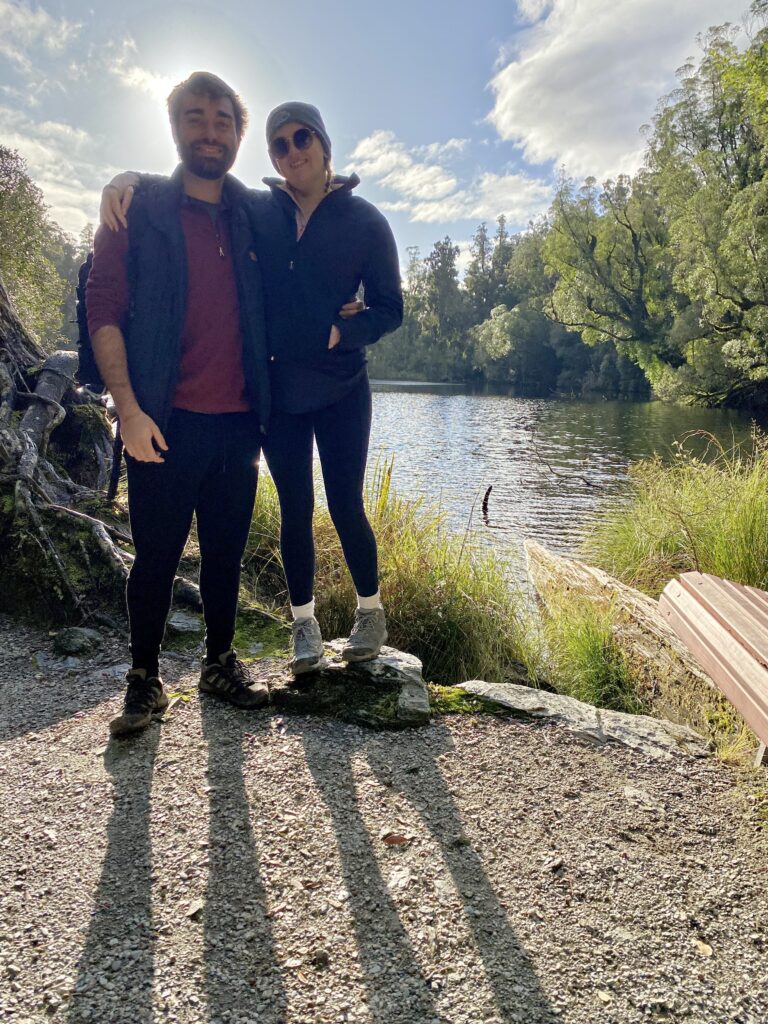 Two people stood in front of Lake Wombat, Franz Josef. The lighting is poor, and their heads are blocking out the sun and you can see their shadows extending from their feet in the direction of the camera.