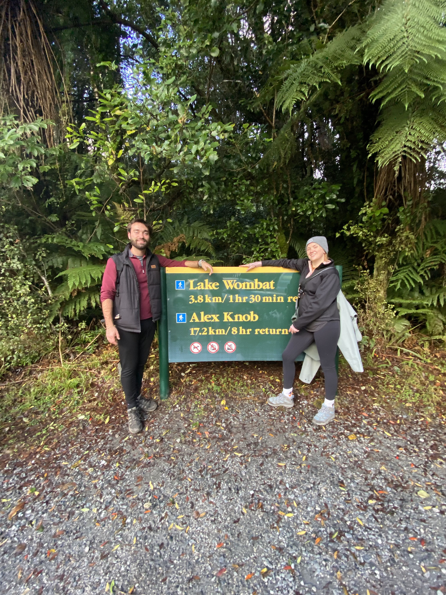 Two people facing forwards, with a sign for the Alex Knob Track and Lake Wombat between them. In the background there is a copious amount of lush forest.