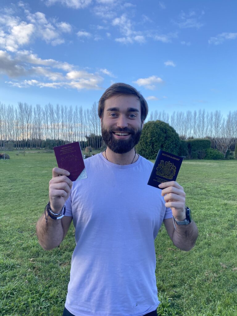 A photo of a man (Ben) holding up both of his passports- his old one and his new one. The background is cloudy blue sky, with a long row of trees in the distance.