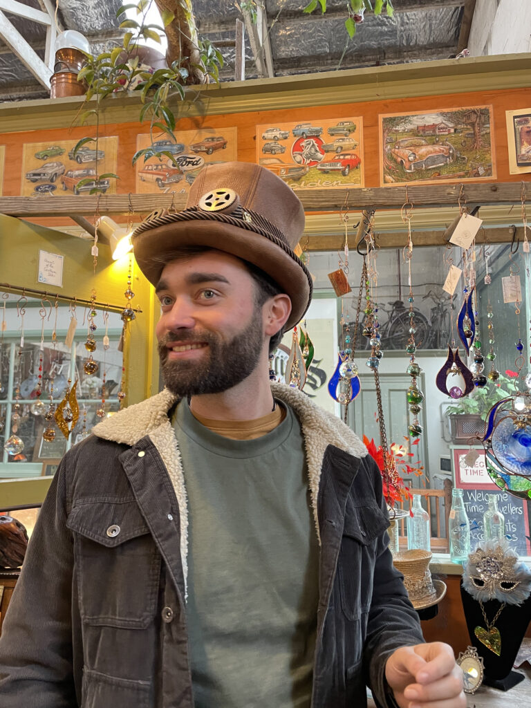 A photo of a bearded man trying on a steampunk style top hat. The hat is brown and has a golden cog on the front.