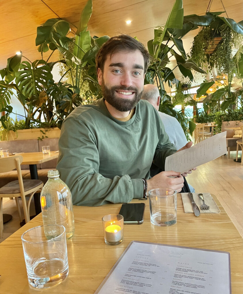 a man with dark hair and a beard wearing a green jumper. sat at a table holding a menu with plants all in the background as decoration in the restaurant