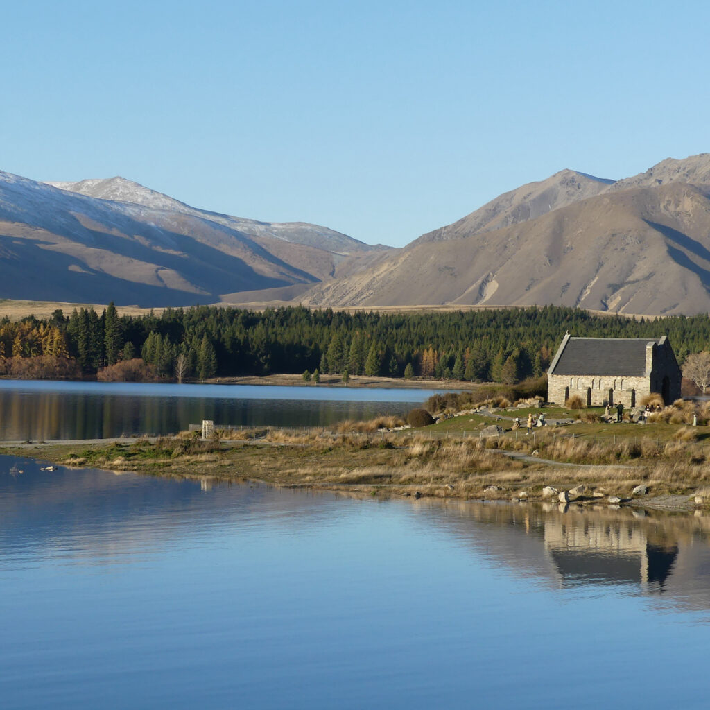 A shot of The Church of the Good Shepherd at golden hour. On the right is the church itself, and the majority of the frame shows the Lake Tekapo and it's surrounding hills.