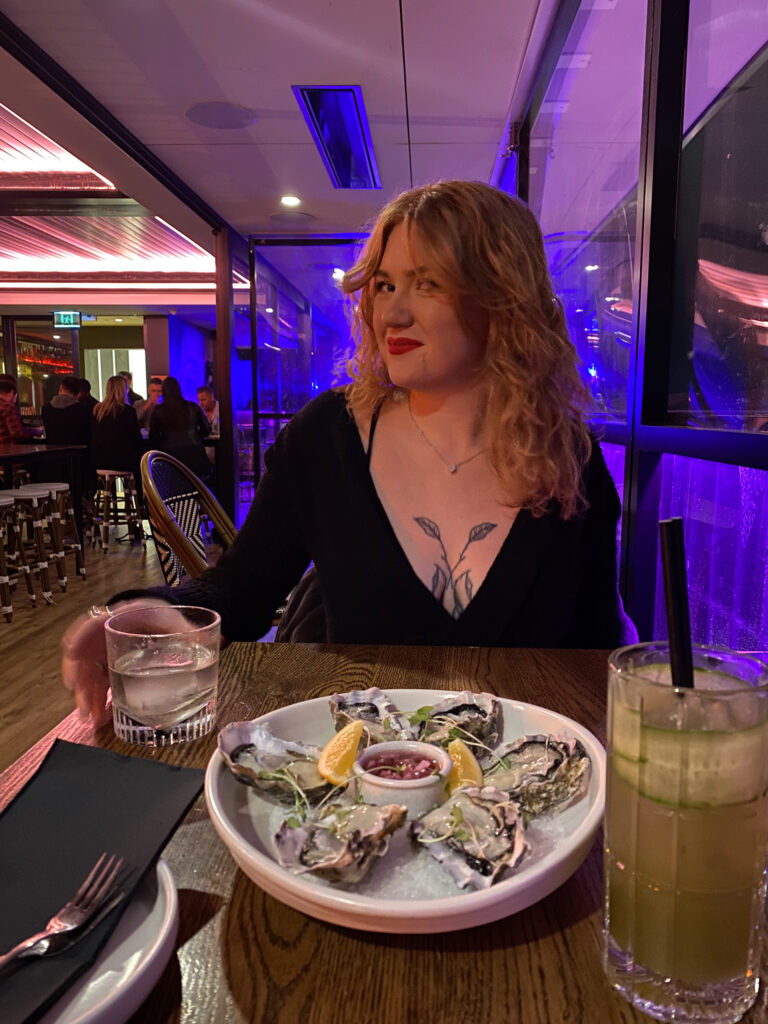 A photo of a pretty, curly haired, smiling young lady who has her hand around a short glass. In front of her is a shallow bowl with six oysters in.