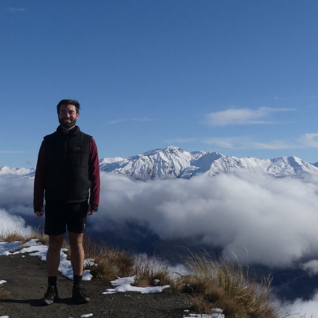 A photo of Ben standing in winter hiking gear at the top of Roys Peak Track (New Zealand), with snowy mountains in the background.