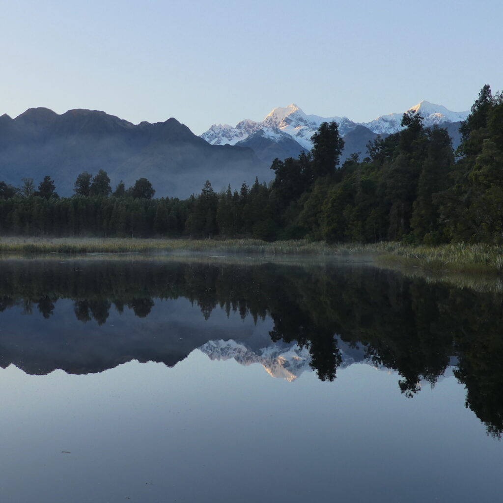 A picturesque Lake Matheson at sunrise. The photo depicts the perfect reflection of Mount Cook and Mount Tasman in the black, still lake.