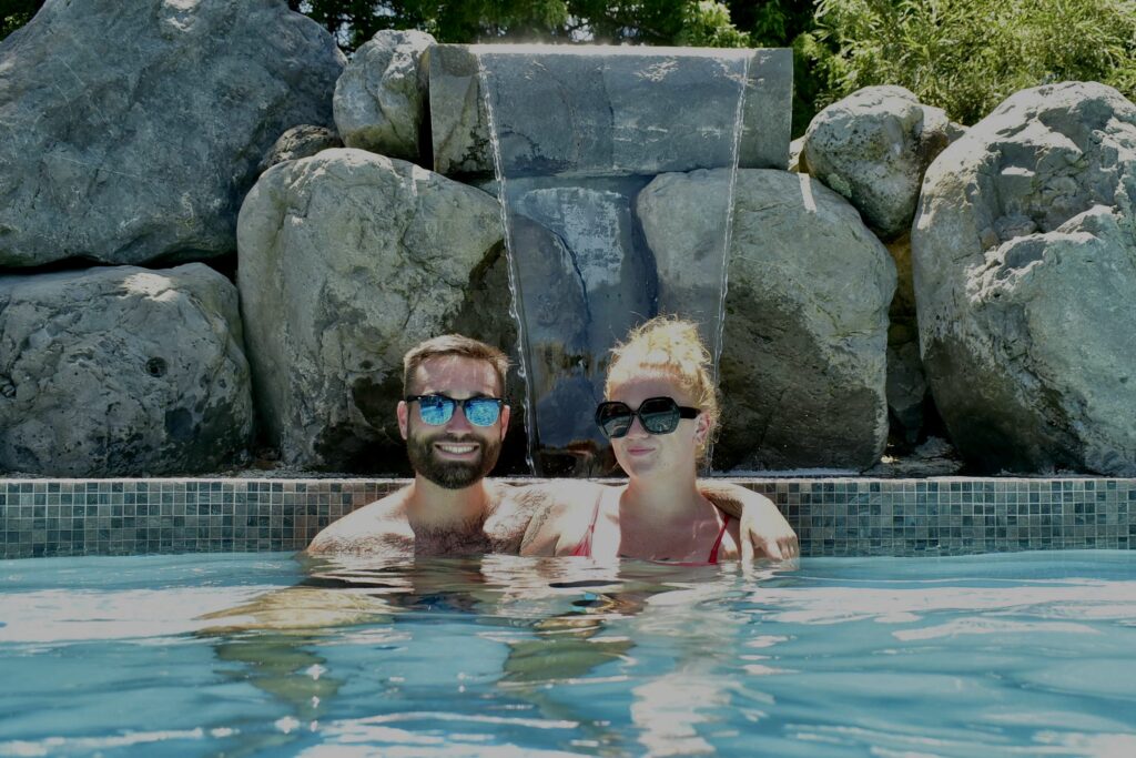 A couple sat in front of a waterfall in the Hanmer Springs Thermal Pool and Spa. Both people are wearing sunglasses and the sun is shining.