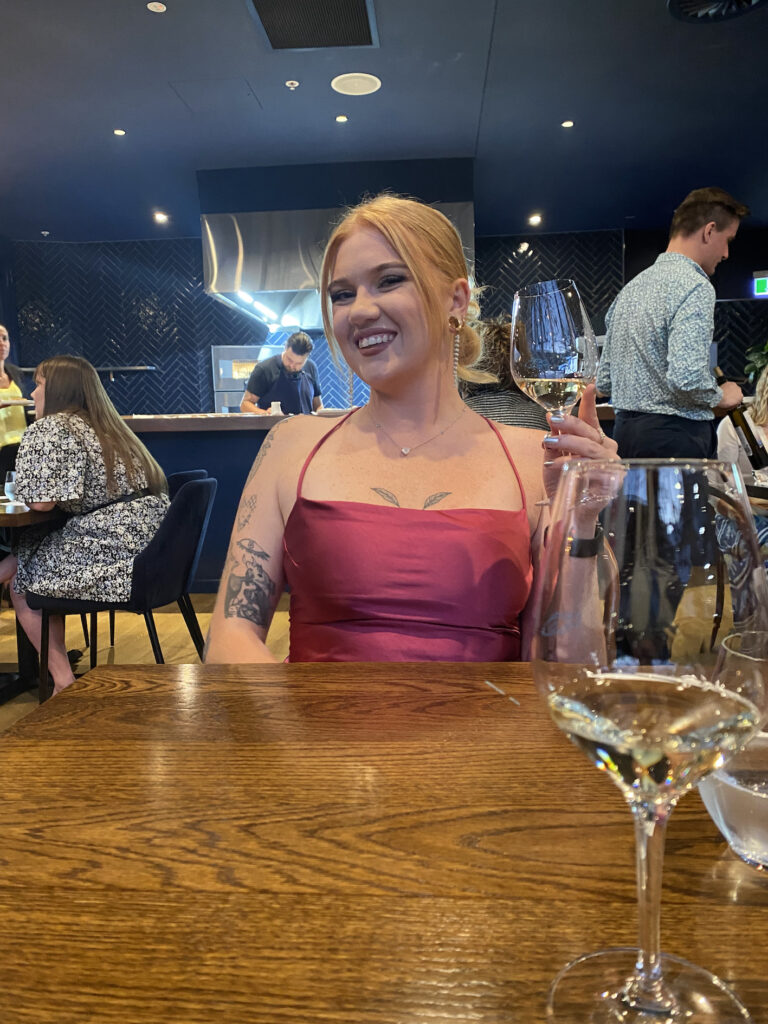 The main feature of this photo is Jess in a stunning pink dress, sat opposite the photographer at a table. There are two glasses of wine visible on the table. We are eating out at a restaurant called Hali, in Christchurch, New Zealand.