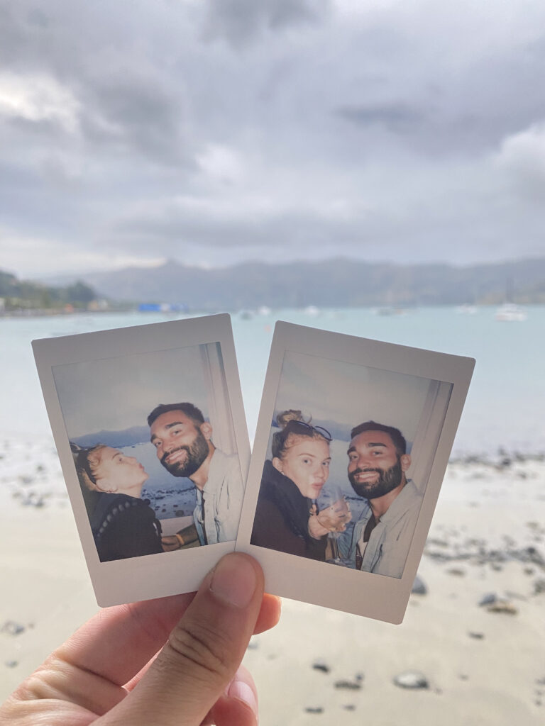 A picture of two photographs taken on an Instax camera, with the beach of Akaroa in the background. The two pictures are a selfie of Ben and Jess, also in front of the beach. Both pictures are similar but different.