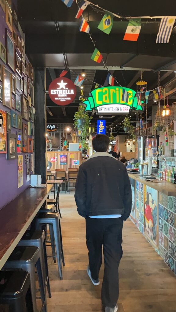 This image shows a man walking through Little High Eatery, Christchurch (New Zealand). There are lots of colourful signs in the background, and a Mexican bar on the right.