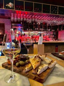 Depicting a glass of wine, a sharing platter with olives, focaccia, and dips, this picture oozes class as it is sat in front of a classy looking wine bar.