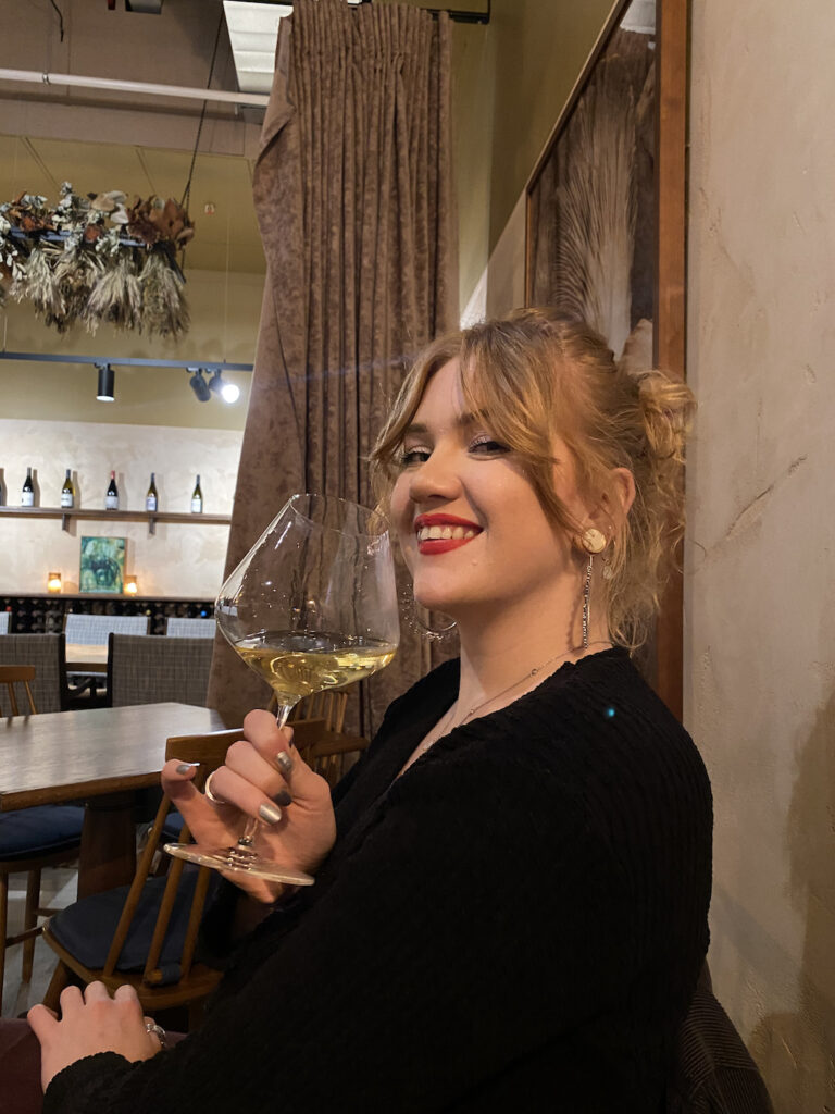 A photo of a beautiful lady, smiling at the camera, holding a glass of white wine.