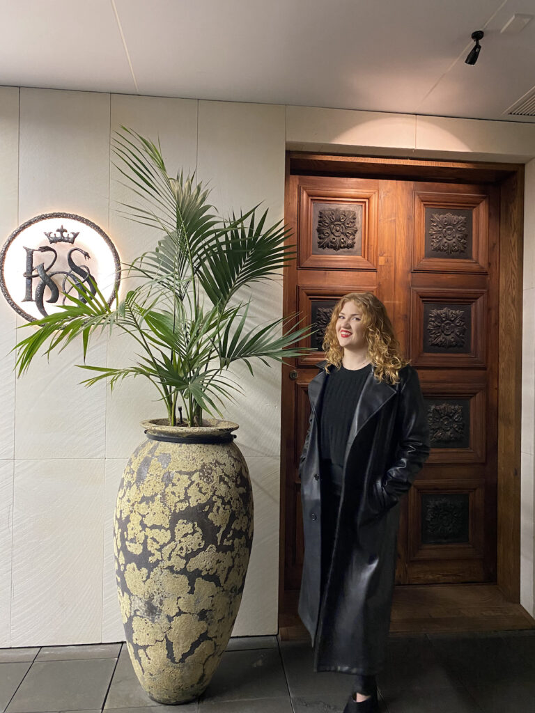 The King of Snake door: a fantastic asian-fusion restaurant in Central Christchurch, New Zealand. In front of the door is a lady wearing a long black leather coat, stood next to a tall green plant, with the King of Snake logo adjacent.