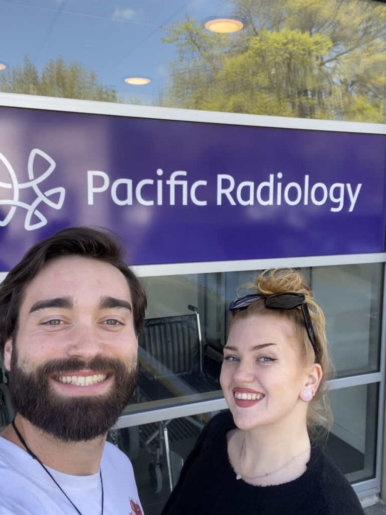 A selfie of a mid-20s couple in front of a "Pacific Radiology" sign. Both people are smiling and look happy; you can see sunglasses on their heads as it is a sunny day. They are at a radiology unit to extend their New Zealand Working Holiday Visas.