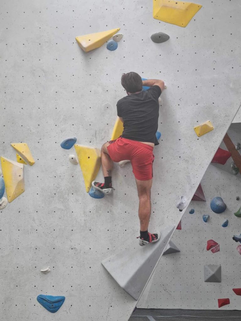 In this photo we can see a man from behind, attempting to climb up a vertical bouldering wall. He is wearing red shorts and a black gym top.