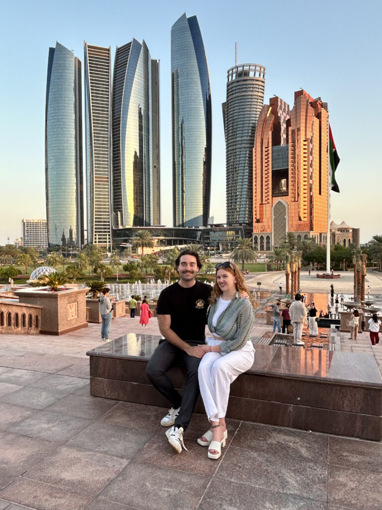 A photo of a couple sitting on a ledge with Emirates Tower (Abu Dhabi, UAE) in the background. The time of day is just before sunset, and the colours are golden.