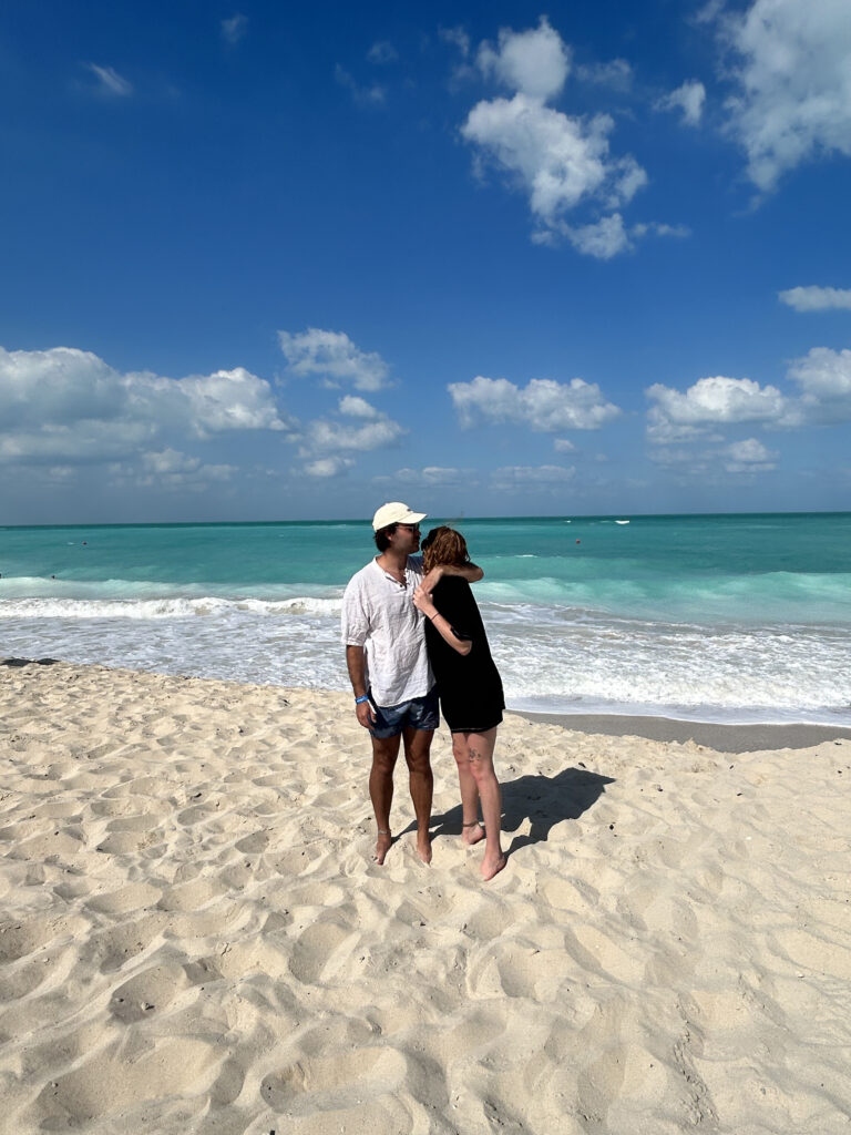 Jess & Ben stood on a beautiful white sandy beach with turquoise ocean sea in the background. Taken in Abu Dhabi, 2023