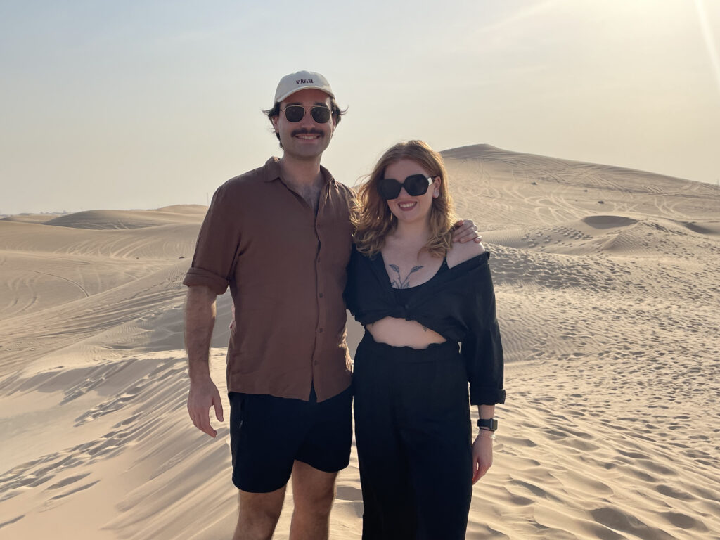 Ben (left) and Jess (right) stood in the desert- you can see miles of golden sand behind them. This was taken in Abu Dhabi, 2023.