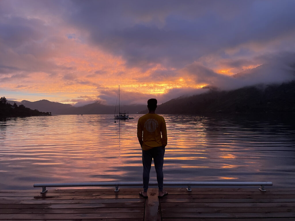 A beautiful sunset, showing orange and light purple colours reflecting on the water at the end of a jetty. This photo was taken in the Endeavour Inlet, Marlborough Sounds, New Zealand.