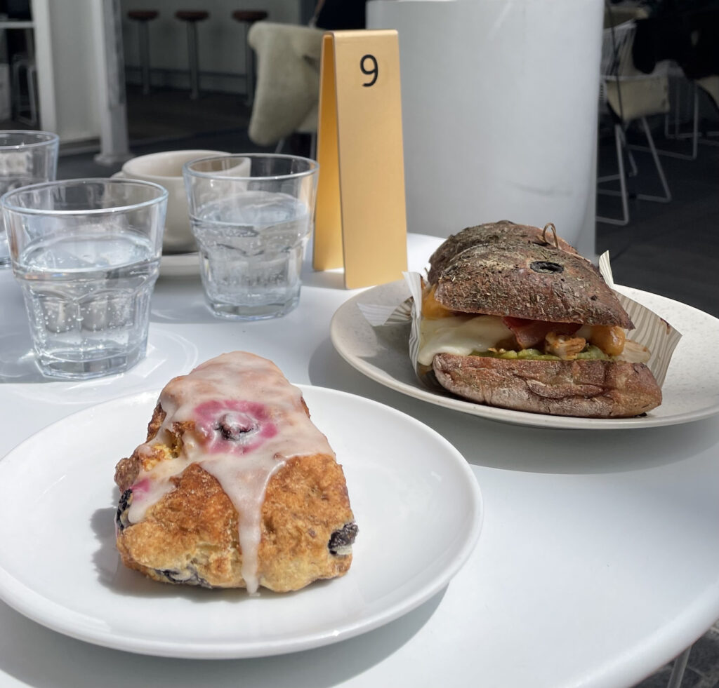 a triangular shaped scone with pink and white icing and a huge sandwich full of avocado and chicken sat on a table in an all whit outdoor seating area of a cafe