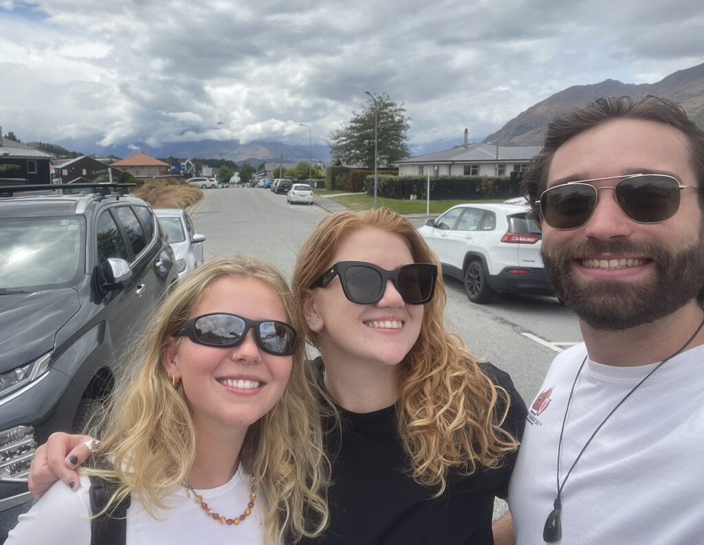 3 friends (2 girls with curly hair and a guy with dark hair and a beard) all stood close together smiling with some moody clouds in the background.