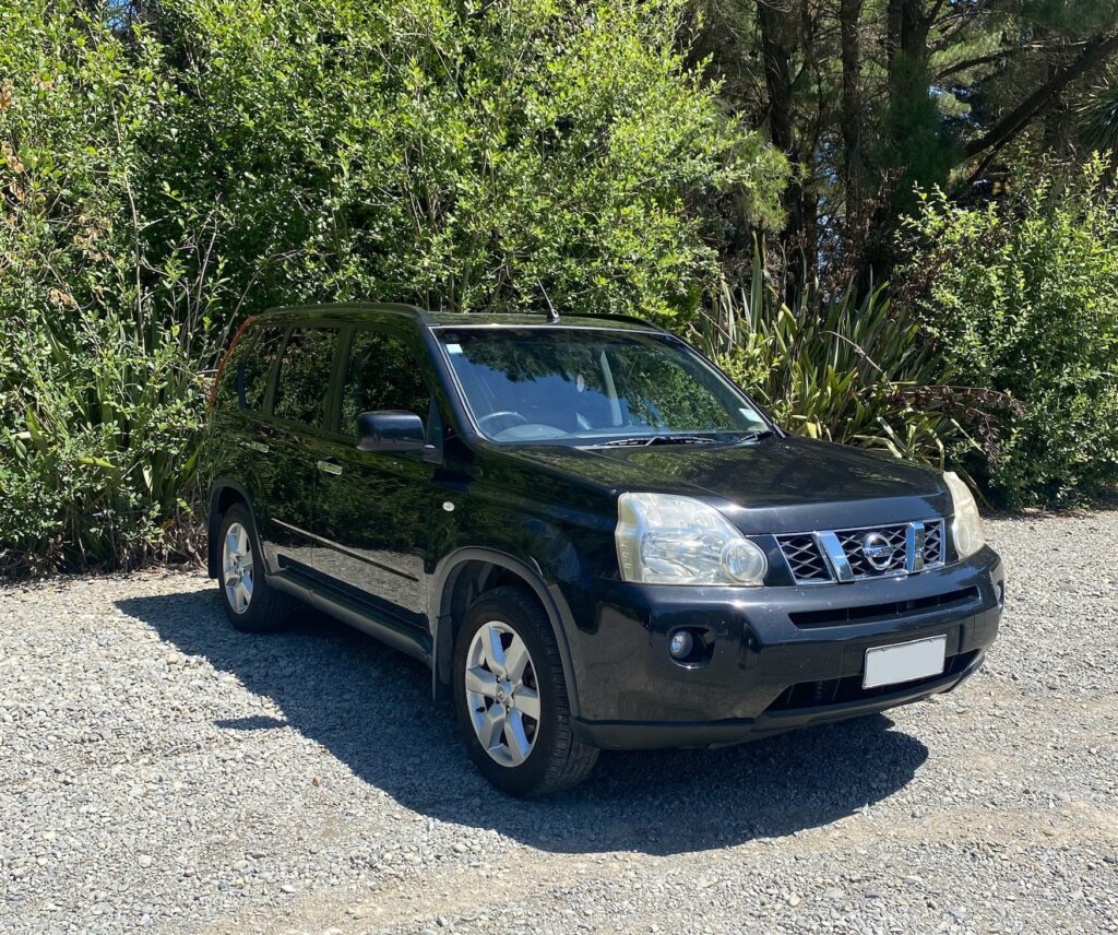 A big black SUV Nissan x trail; parked in front of some green bushes/trees on a sunny day.