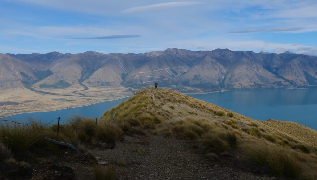 In the middle picture is a man stood on a grassy peak with a bright blue lake below him on either side and in the background is a row of sunlit mountain tops