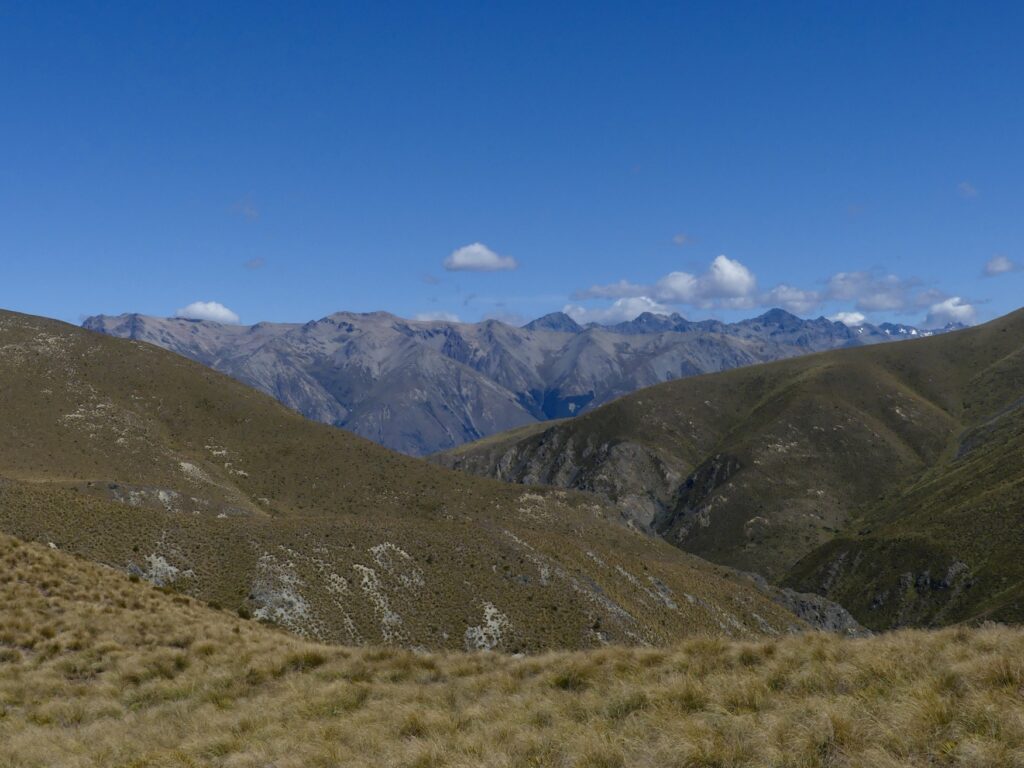 a photo with four different layers to the ground, in the foreground in a grassy patch that is being walked on, in the front mid ground there are some drier grassy hills. Then the far mid ground, is a row of rocky mountains with a clear blue sky over head in the background.