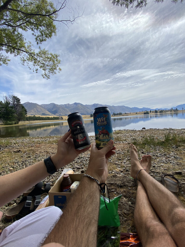 A picture depicting a picnic set up on Lake Middleton, New Zealand. You can see two Emerson's craft beers being held up.