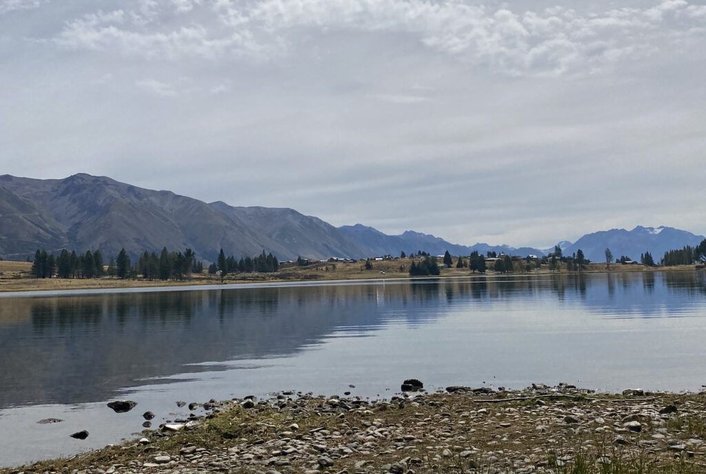 A photo of Lake Middleton, near Lake Ohau in the Mackenzie District of New Zealand. We can see the lake and the grey, stoney mountains in the background. The lake is reflecting the image in it's still waters.