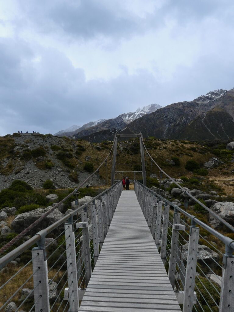 a wooden path onto a bridge over the Hooker Valley river with mountains in the background on a cloudy day