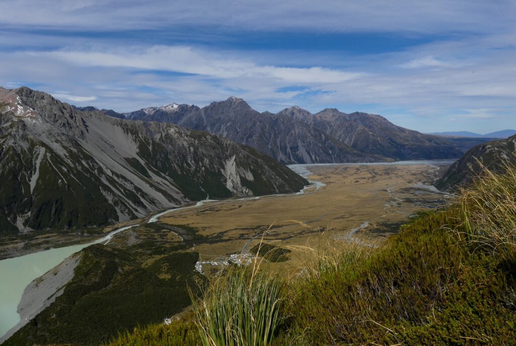 the view over mount cook village valley with hills and mountains in the background with a river flowing through the grassy grounds.