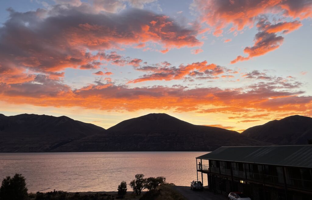 A beautiful sunrise over Ben Ōhau and Lake Ōhau taken from Lake Ōhau Lodge. The colours in the sky are red and orange with the light blue sky behind. A silhouette of the mountains and the lodge can be seen.