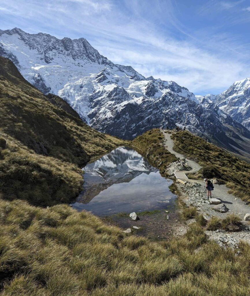 A photo of Sealy Tarn along the Mueller Hut route in Mount Cook National Park. In the background you can see the Southern Alps covered with a light dusting of snow. The weather is good, blue skies with soft clouds.