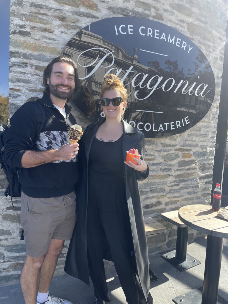 A photo of Jess and Ben from "Jess and Ben Travel" in front of the sign for Patagonia Ice Creamery in Queenstown. Both are holding a cold treat from the store.