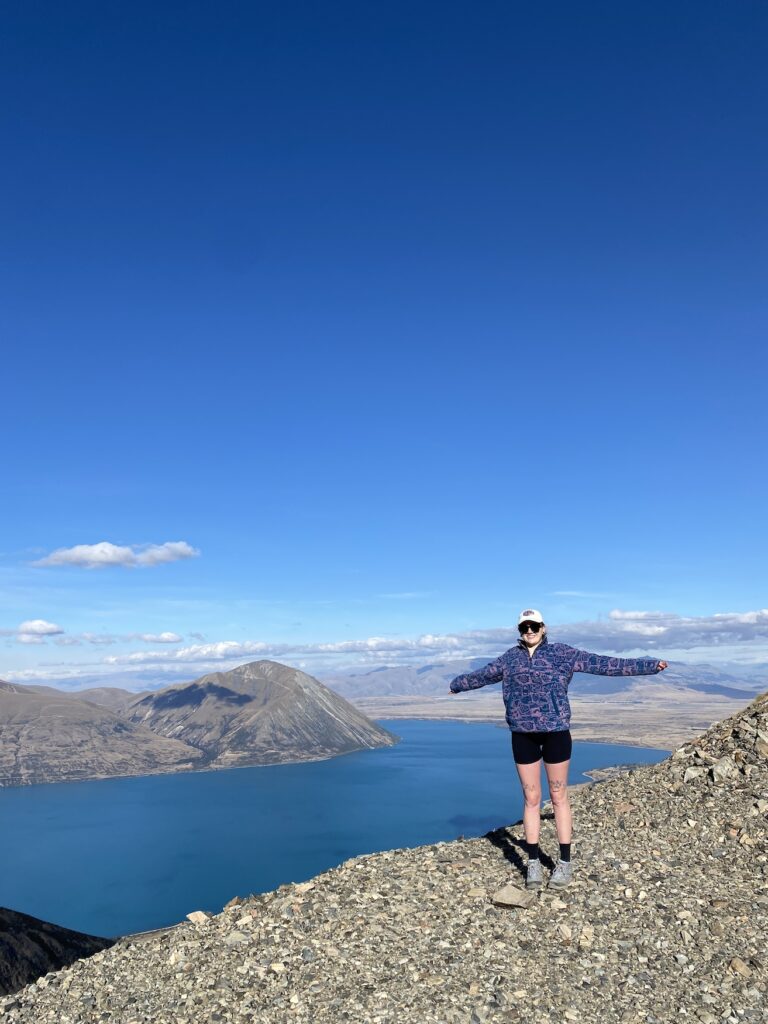 Jess from "Jess and Ben Travel" stood on the edge of a path with her arms outstretched. The magnificent view behind her is of Lake Ōhau, which is a deep blue in colour, and Ben Ōhau, the highest peak in the immediate area.