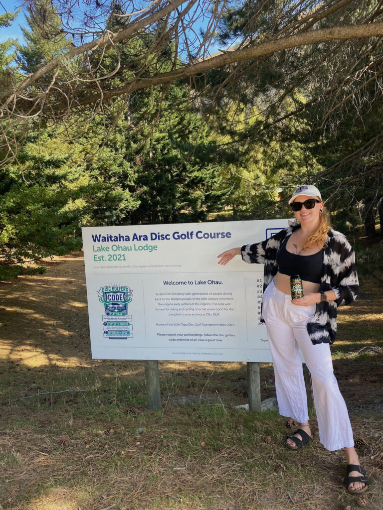 A photo of Jess from "Jess and Ben Travel" posing by the Waitaha Art Disc Golf Course at Lake Ōhau Lodge. She is holding an Emersons beer.