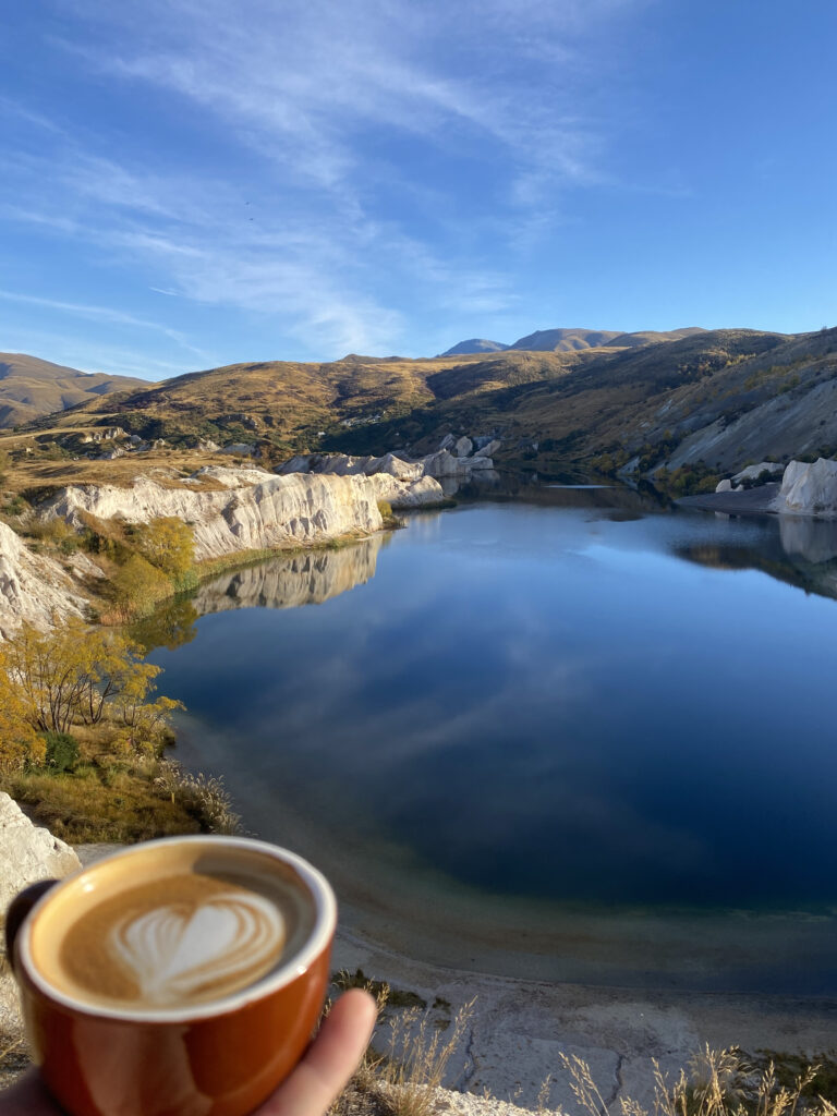 The Blue Lake, Saint Bathans. This picture is looking out onto the lake in the morning, and the photographer is holding a cup of coffee.